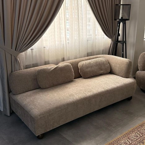 کاناپه سهنفره پارچهای کرم با طراحی ساده و کوسنهای پشتی، قرارگرفته جلوی پنجره بزرگ با پردههای طوسی و یک آباژور ایستاده در گوشه اتاق.